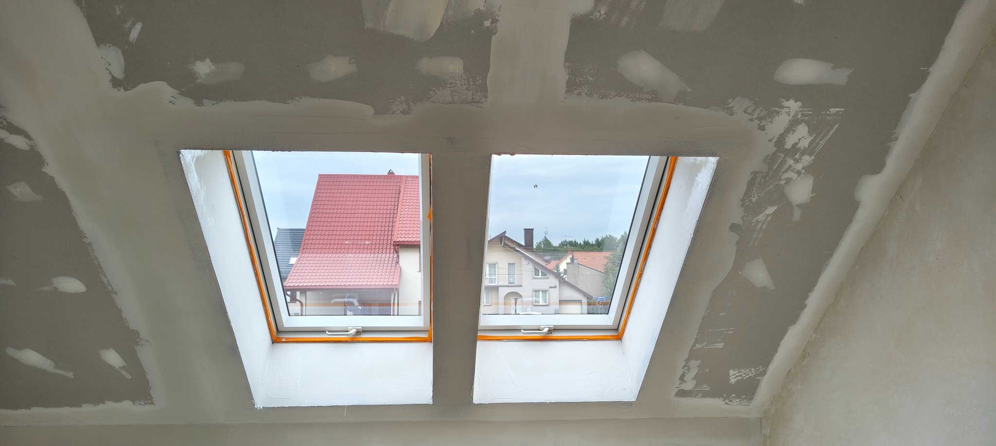 Poddasze z dwoma oknami dachowymi po szpachlowaniu, widoczne ślady gładzi na ścianach i suficie. W oknach widok na domy z czerwonymi dachami.