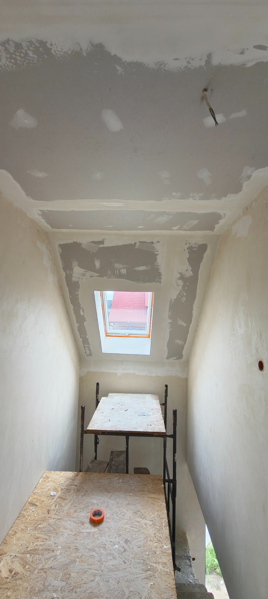 Poddasze w trakcie szpachlowania z oknem dachowym i rusztowaniem. Widoczne niedoskonałości, ślady po łatach. Pomarańczowa taśma na rusztowaniu.
