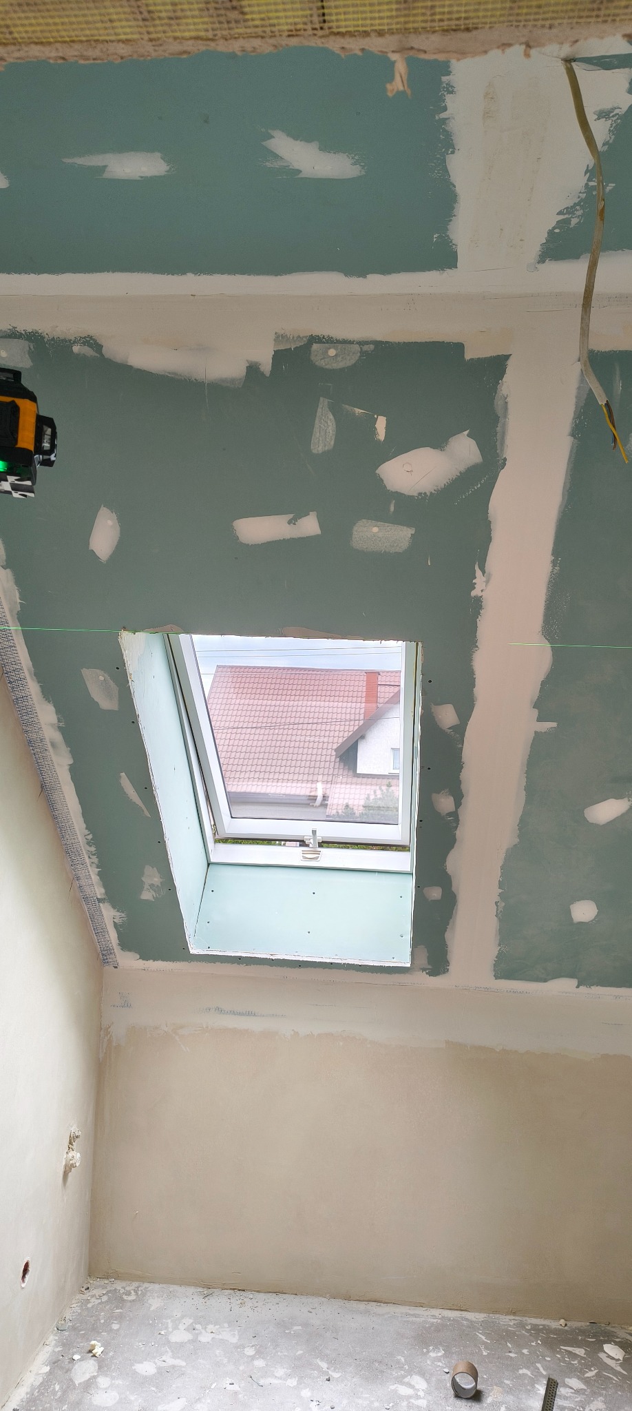 Montaż płyty KG na poddaszu z oknem dachowym. Widoczne szpachlowane łączenia, laser krzyżowy i fragment siatki na suficie. Surowy stan wykończenia wnętrza.