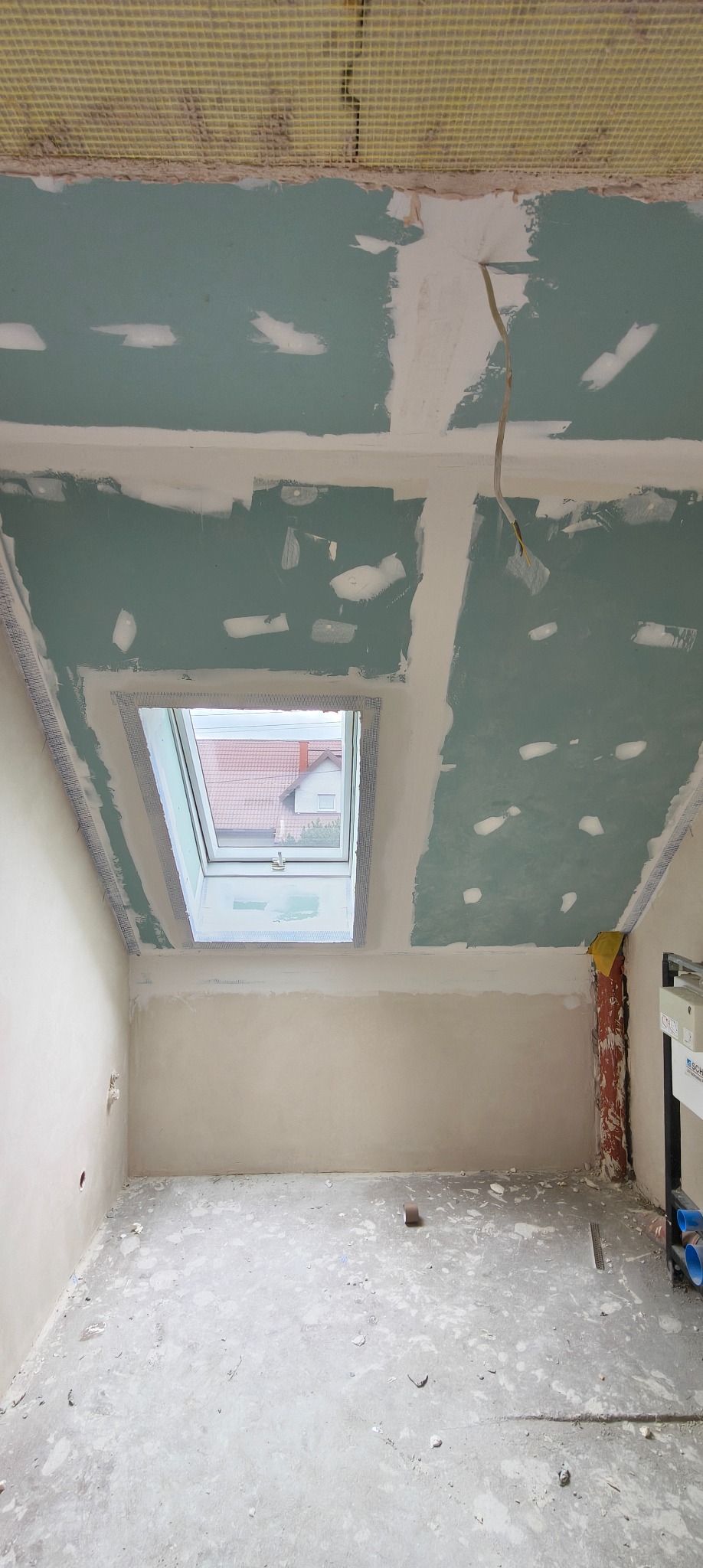 Pokój na poddaszu w trakcie prac wykończeniowych, widoczne płyty gipsowo-kartonowe na suficie z oknem dachowym i częściowo wyszpachlowane ściany.