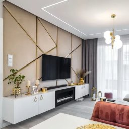 FINE-DESIGN SPÓŁKA CYWILNA PAWEŁ LACHOWSKI, GRZEGORZ SIWIK - Elegancki salon z beżową ścianą zdobioną złotymi liniami, białymi szafkami RTV z paleniskiem i subtelnym oświetleniem. Nowoczesny design wnętrza.