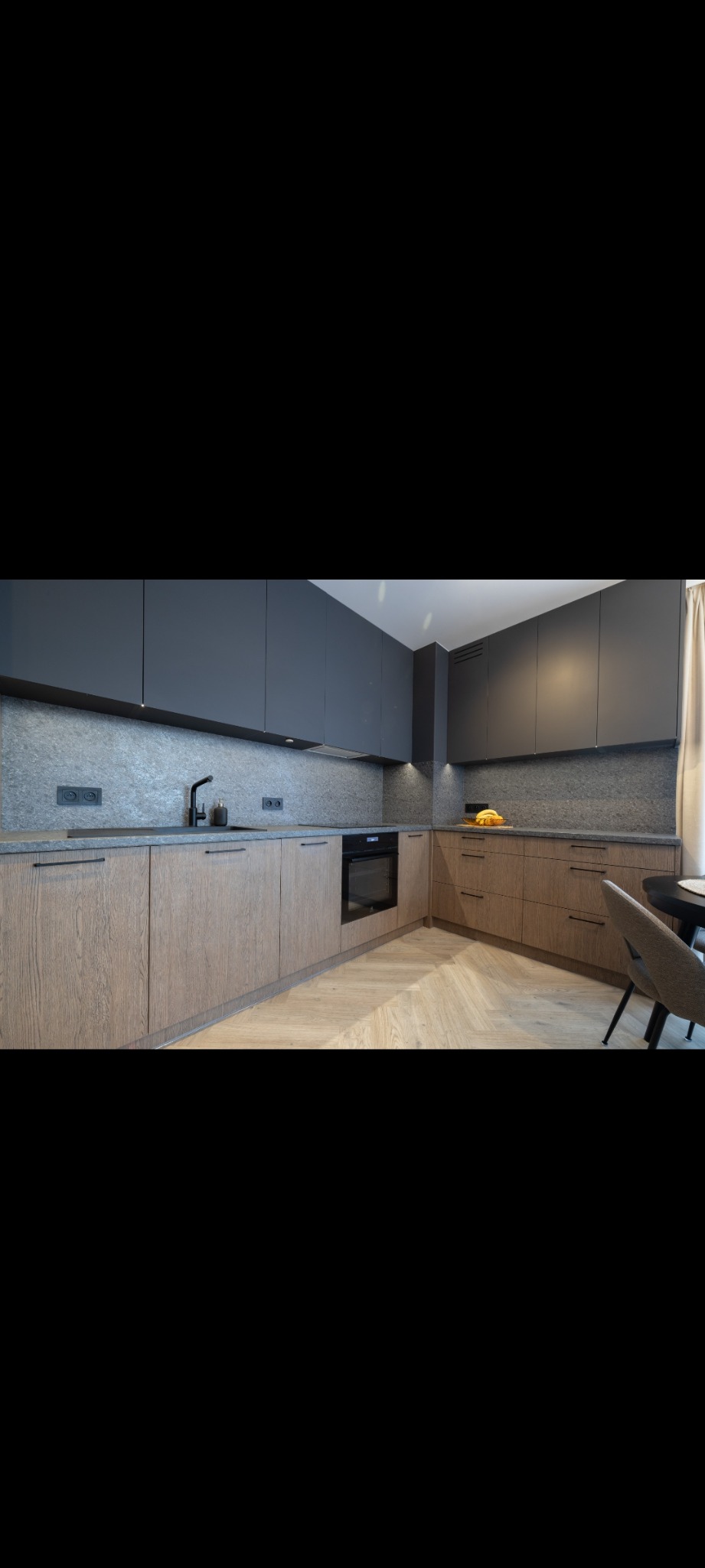 Nowoczesna kuchnia z szarymi górnymi szafkami i drewnianymi dolnymi, blat w kolorze kamienia, czarna armatura i parkiet w jodełkę. Minimalistyczny design.