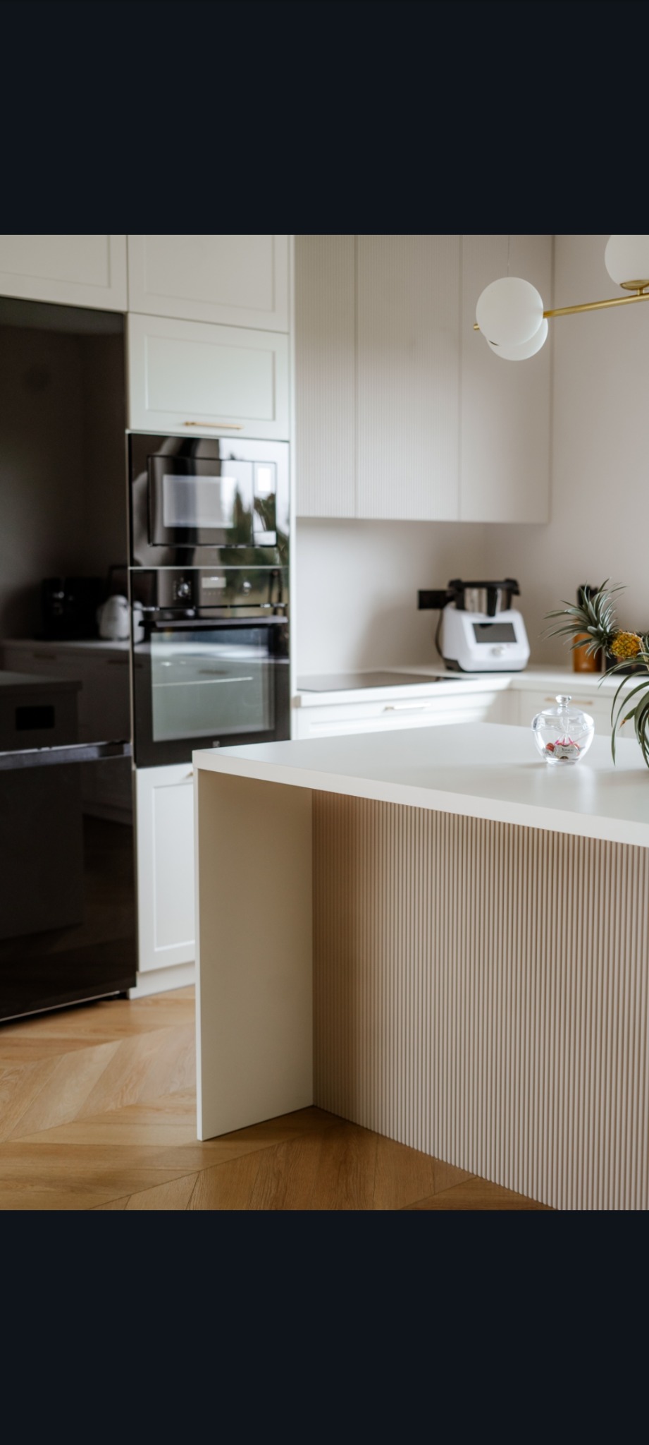 Jasna kuchnia z wyspą i ryflowanymi frontami, oświetlona lampą kulistą. Widoczny sprzęt AGD i parkiet w jodełkę.