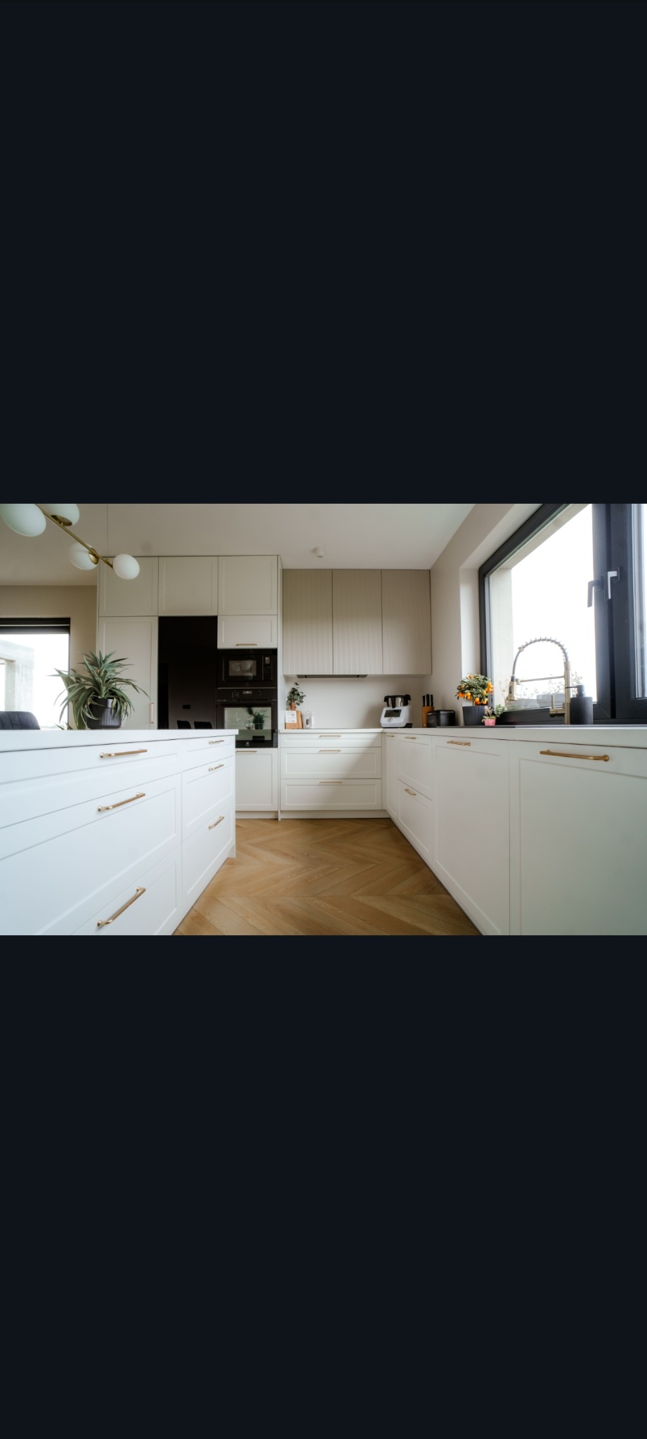 Jasna kuchnia z wyspą, fronty szafek białe z poziomymi uchwytami. Na blacie kwiaty i akcesoria kuchenne. Podłoga z parkietu ułożonego w jodełkę.