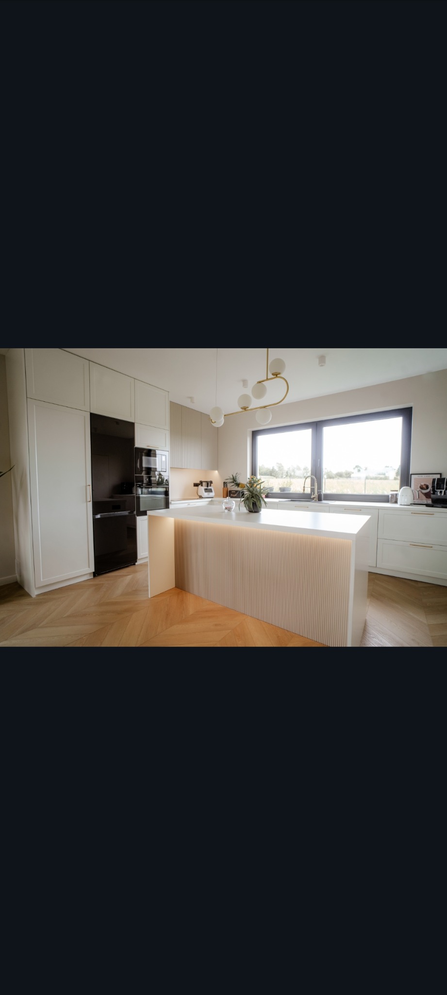 Jasna kuchnia z wyspą oświetloną ledami, parkiet w jodełkę, czarna lodówka i duże okno z widokiem na zieleń. Nowoczesny design, ciepłe światło.