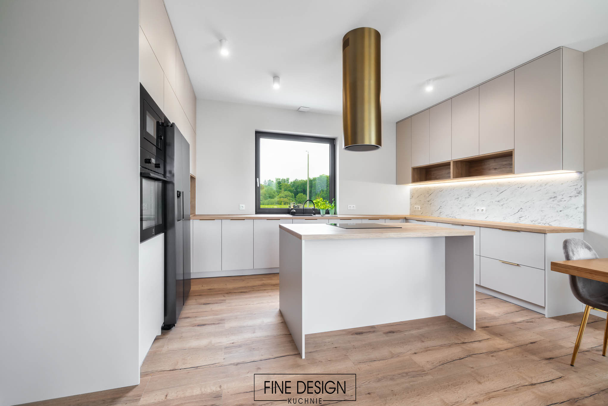 Nowoczesna kuchnia z wyspą, białe szafki z drewnianymi blatami, złoty okrągły okap i parkiet w jodełkę. Minimalistyczny design z widokiem na zieleń.