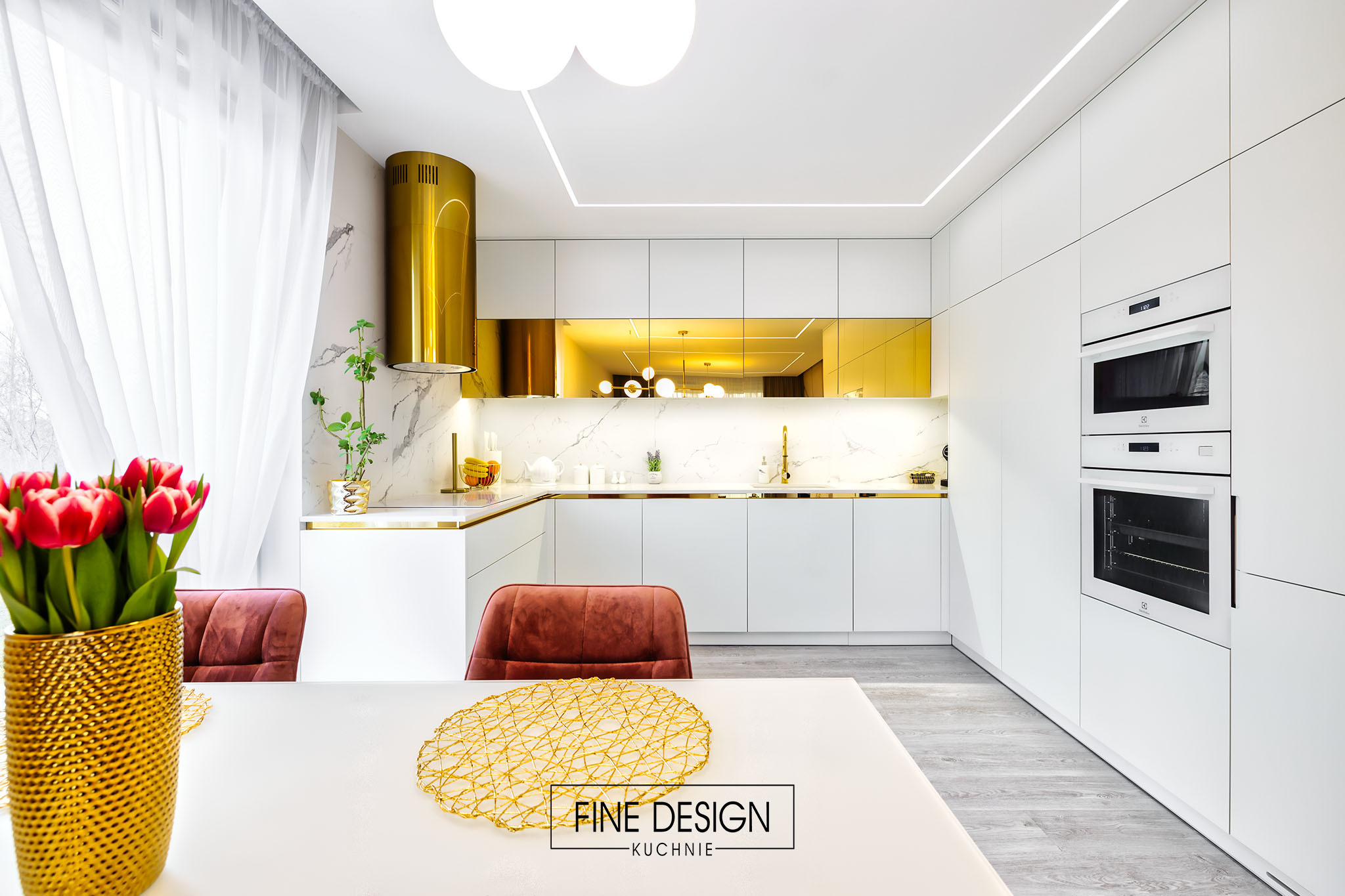 Nowoczesna kuchnia z białymi frontami, złotymi akcentami i marmurowym blatem. Na pierwszym planie stół z bukietem tulipanów i złotą podkładką.
