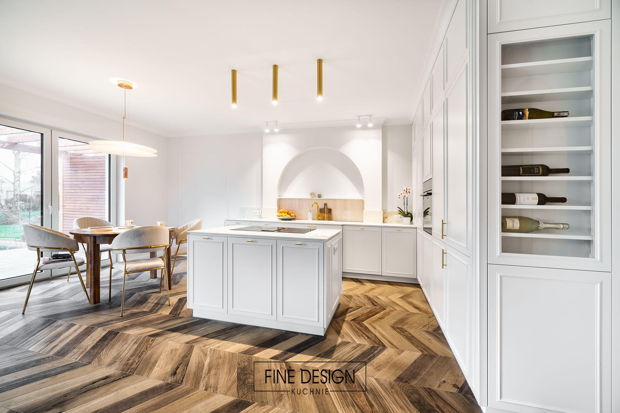 Elegancka, biała kuchnia z wyspą i parkietem w jodełkę. W tle stół z krzesłami i okno. Szafka z winami po prawej. Złote akcenty oświetleniowe.