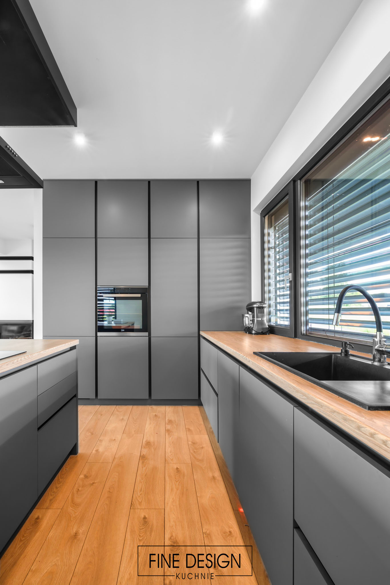 Szara kuchnia na wymiar z drewnianym blatem, widok na okno z żaluzjami i sprzęt AGD. Nowoczesny design z drewnianą podłogą i czarnymi detalami.