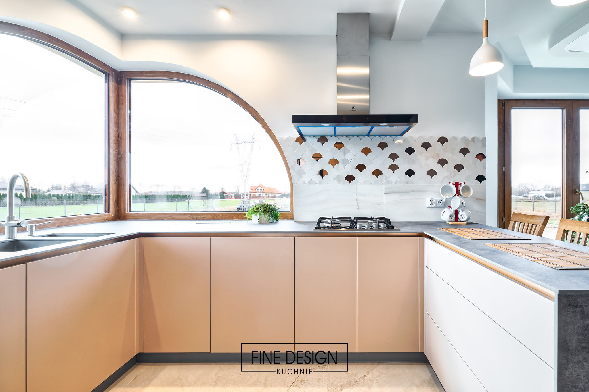 Jasna kuchnia z beżowymi szafkami, dużym oknem łukowym i dekoracyjną mozaiką nad kuchenką. Nowoczesny design z elementami drewna.