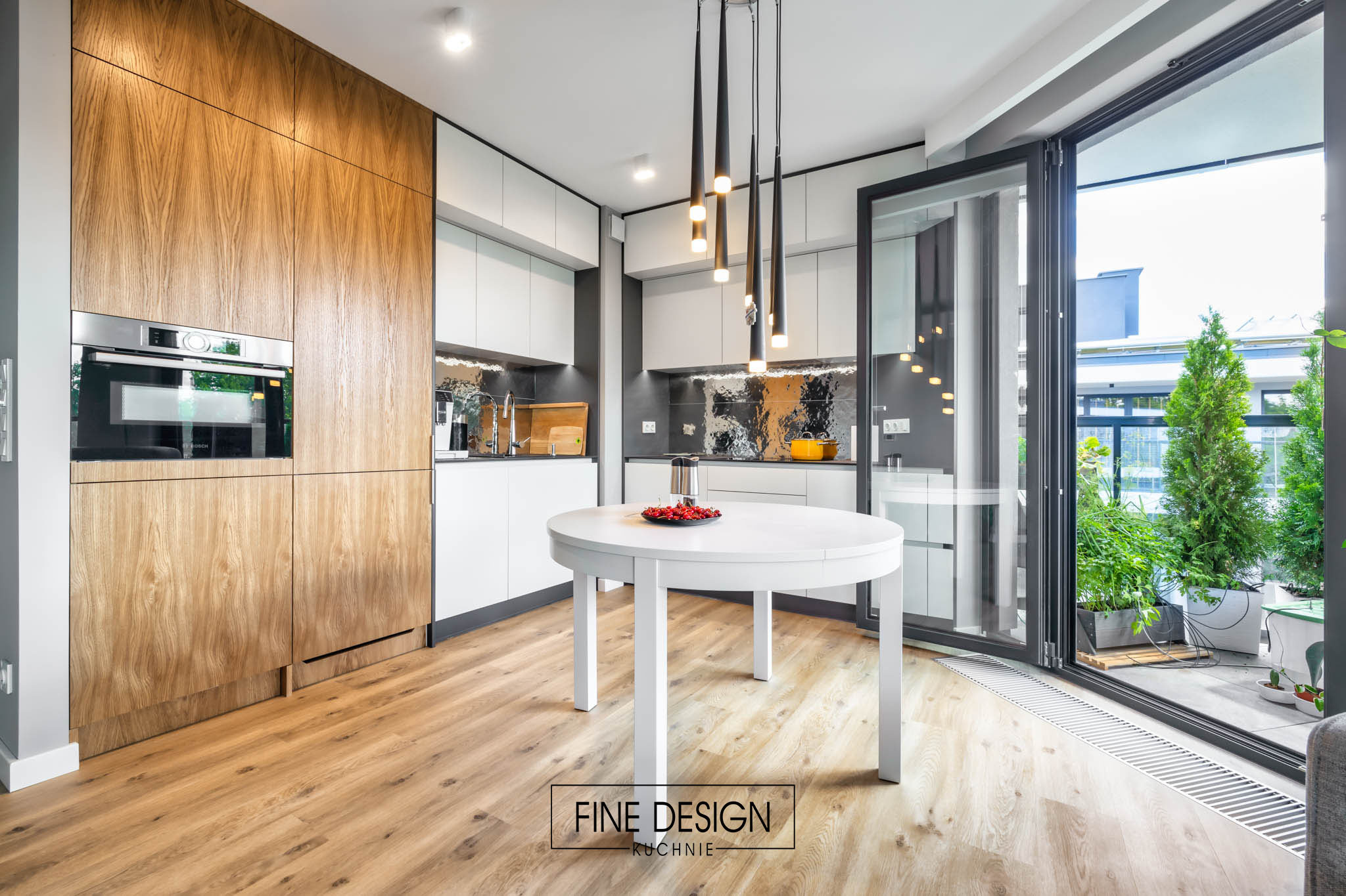 Nowoczesna kuchnia z drewnianą zabudową, białymi szafkami i okrągłym stołem. Widok na balkon przez rozsuwane drzwi. Minimalistyczne lampy wiszące.