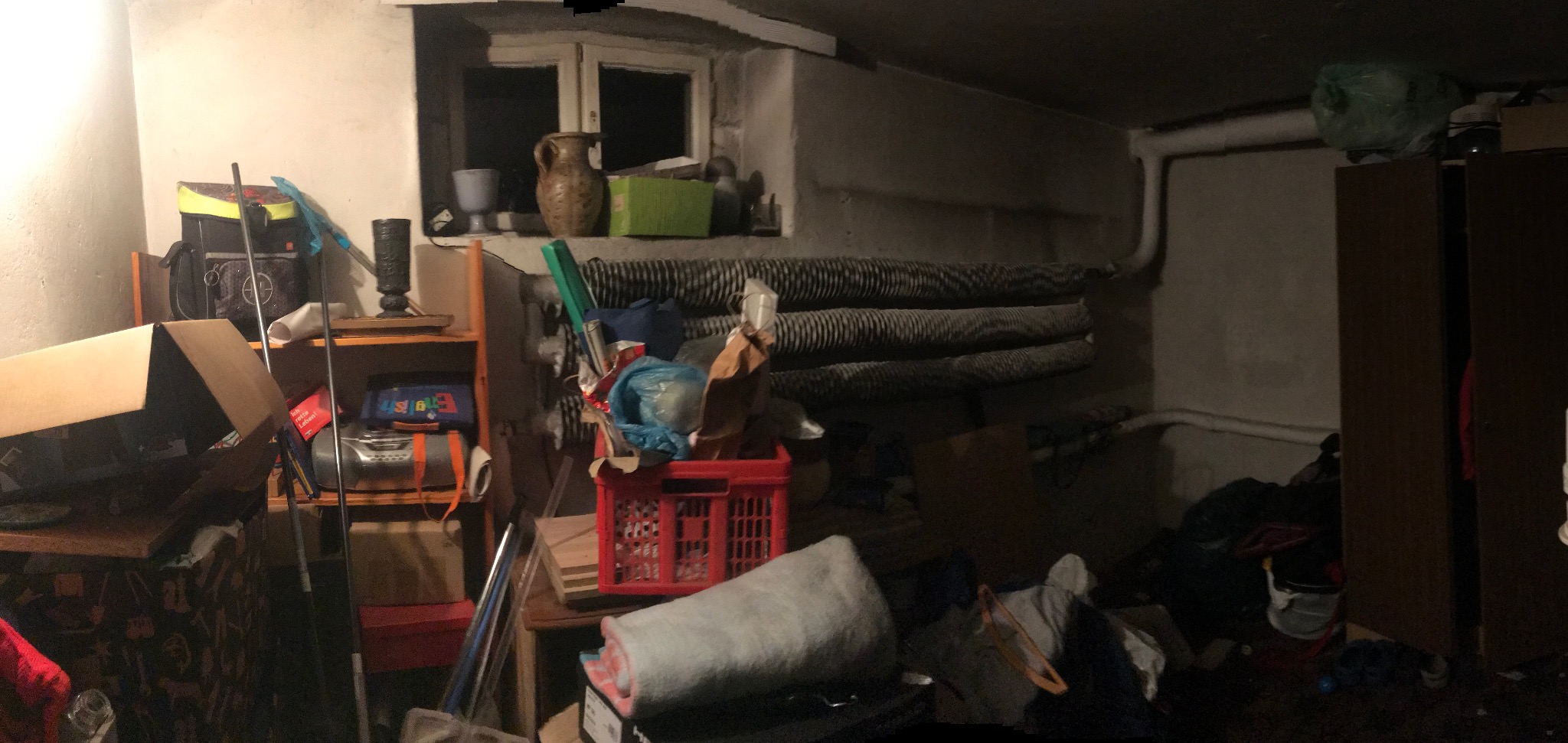 Zagracone pomieszczenie piwniczne z oknem, grzejnikiem i różnymi przedmiotami, w tym kartonami, meblami i skrzynkami. Panuje półmrok.
