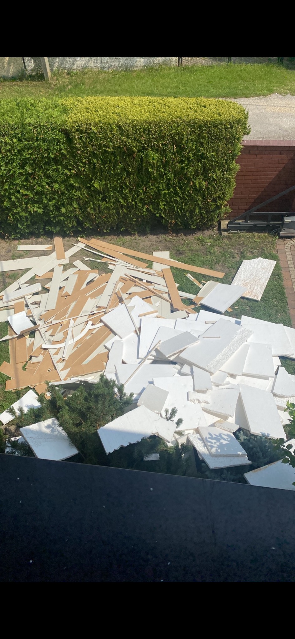 Stos białych i brązowych płyt na trawie przed żywopłotem i ceglanym murem. Gruz po remoncie na zewnątrz.