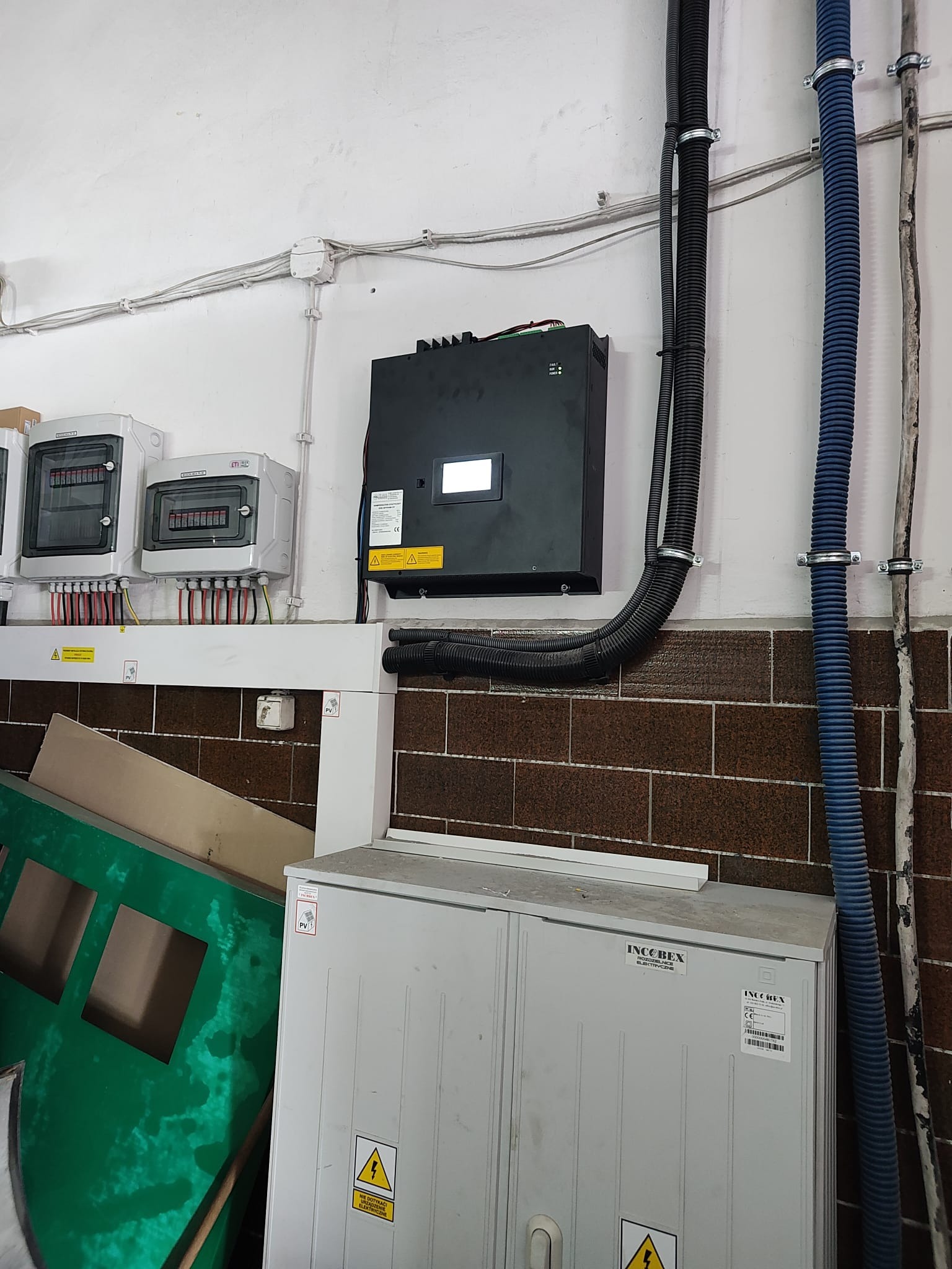 Czarna skrzynka inwertera na ścianie nad szafą elektryczną. Widoczne przewody, kable i oznaczenia ostrzegawcze. Instalacja fotowoltaiczna w pomieszczeniu.