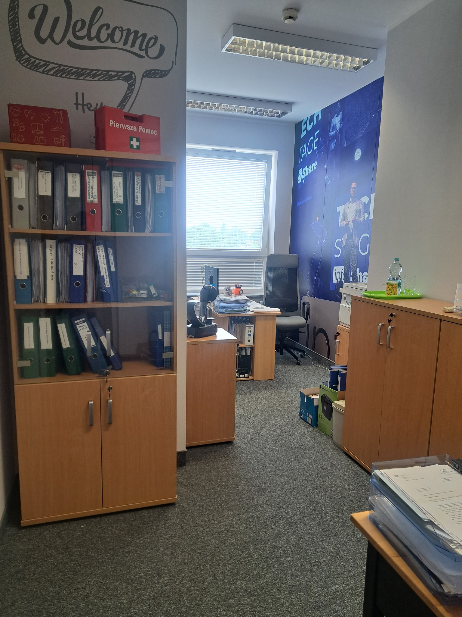 Jasne biuro z regałem pełnym segregatorów, apteczką pierwszej pomocy i plakatem firmowym na ścianie. Widok na biurko i krzesło biurowe. Schludne i zorganizowane.