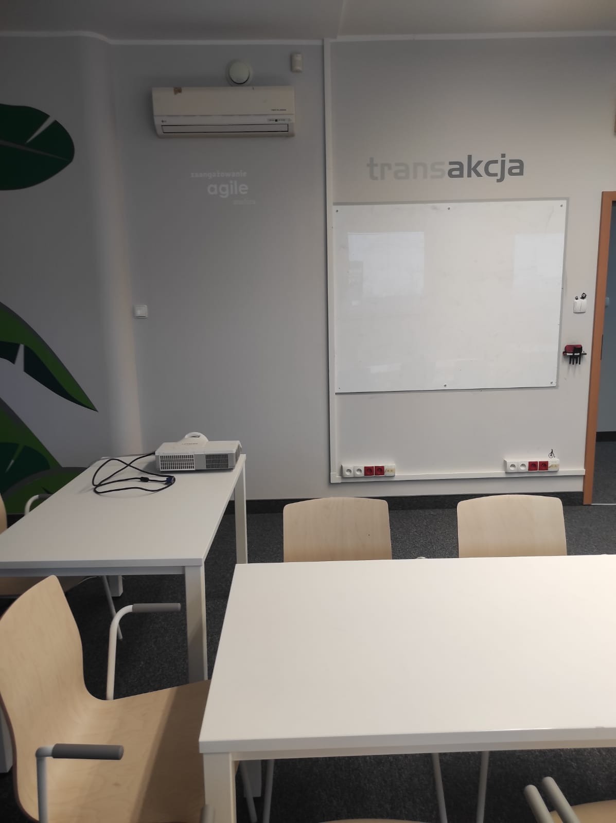 Sala szkoleniowa z projektorem, białymi stołami i krzesłami. Na ścianie tablica suchościeralna z napisem 'transakcja' i hasło 'zaangażowanie agile'.