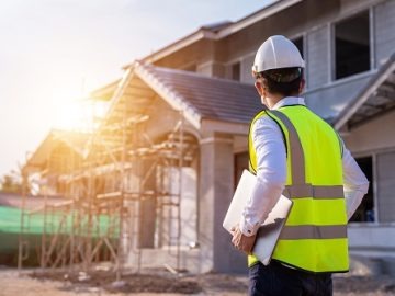 Inspektor BHP w żółtej kamizelce i kasku, z laptopem, kontroluje budowę domu jednorodzinnego w słoneczny dzień. Widoczne rusztowania i stan surowy otwarty.