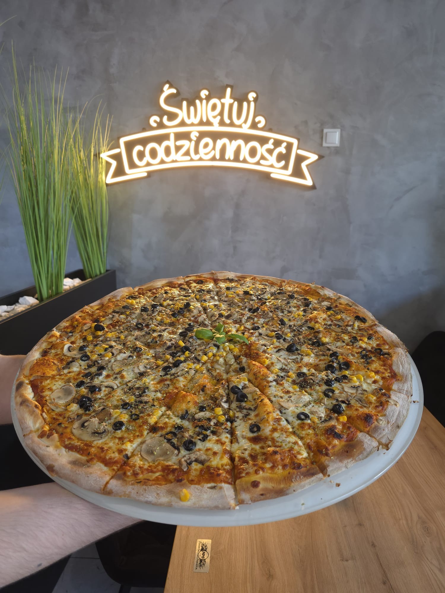 Ogromna pizza z kukurydzą i oliwkami na białym talerzu, serwowana w Starachowicach. W tle neonowy napis 'Świętuj codzienność' i minimalistyczny wystrój.