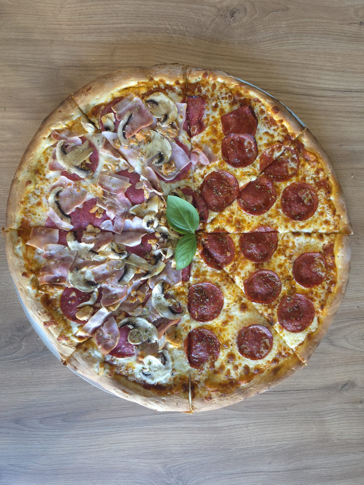 Podwójna pizza na drewnianym stole: połowa z pieczarkami i szynką, druga z pepperoni. Listek bazylii w centrum. Widok z góry, ciepłe światło.