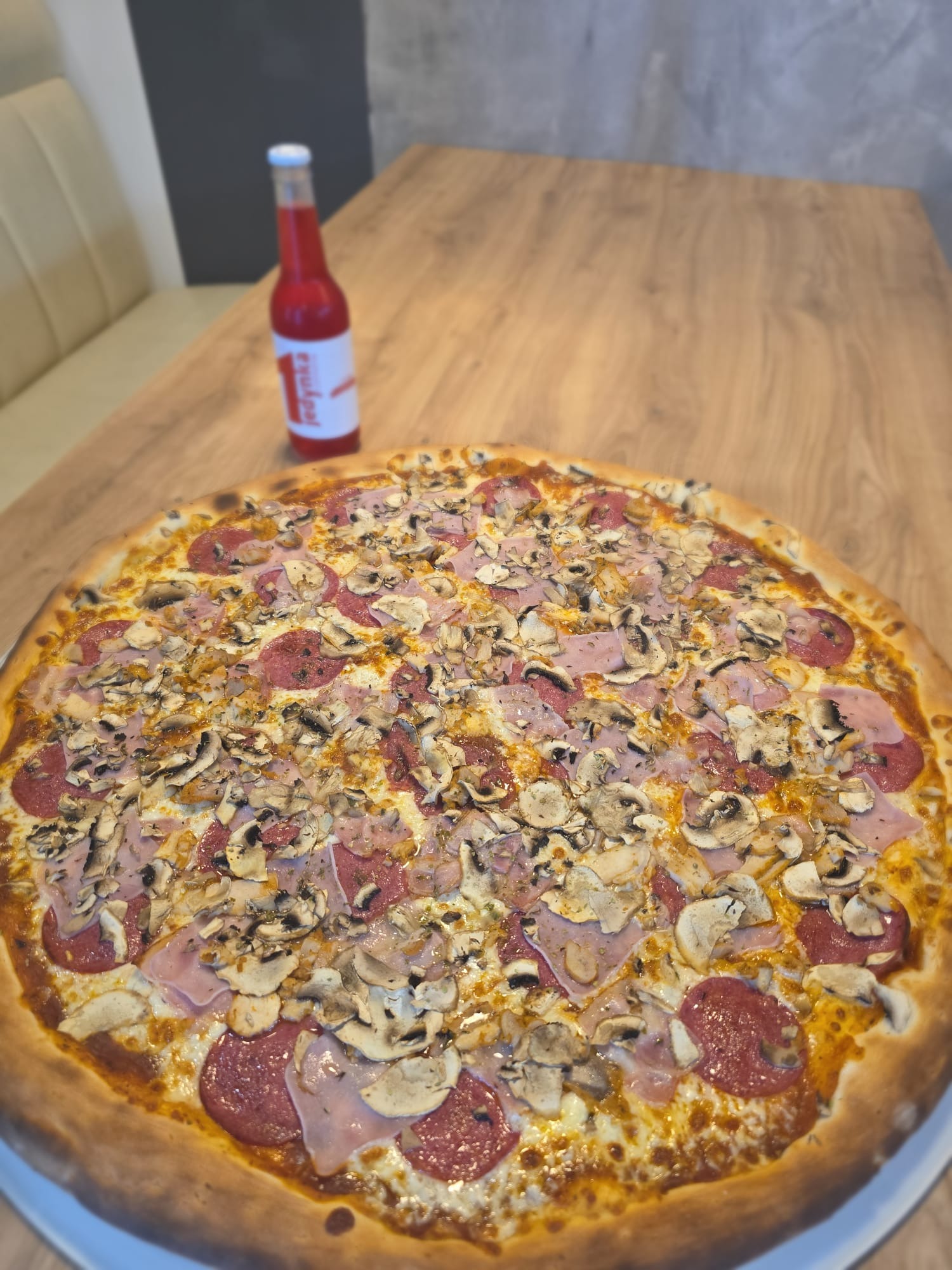 Ogromna pizza z salami, szynką i pieczarkami na drewnianym stole, obok butelka czerwonego napoju. Idealna propozycja na catering.