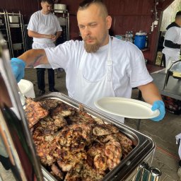 SPÓŁDZIELNIA SOCJALNA MNIEJ WIĘCEJ - Kucharz w niebieskich rękawicach nakłada grillowane mięso na talerz. W tle drugi kucharz i pojemniki z jedzeniem. Catering na świeżym powietrzu, impreza plenerowa.