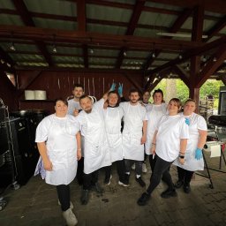 SPÓŁDZIELNIA SOCJALNA MNIEJ WIĘCEJ - Zespół kucharzy w białych fartuchach i koszulkach pozuje na tle drewnianej konstrukcji z oświetleniem. Widoczny sprzęt gastronomiczny i kostka brukowa.