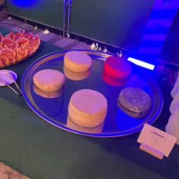 Srebrna taca z selekcją ser&oacute;w (r&oacute;żne rodzaje i kolory) oraz grejpfrut&oacute;w na zielonym obrusie, oświetlona niebieskim światłem, z widoczną etykietą 'cheese'.