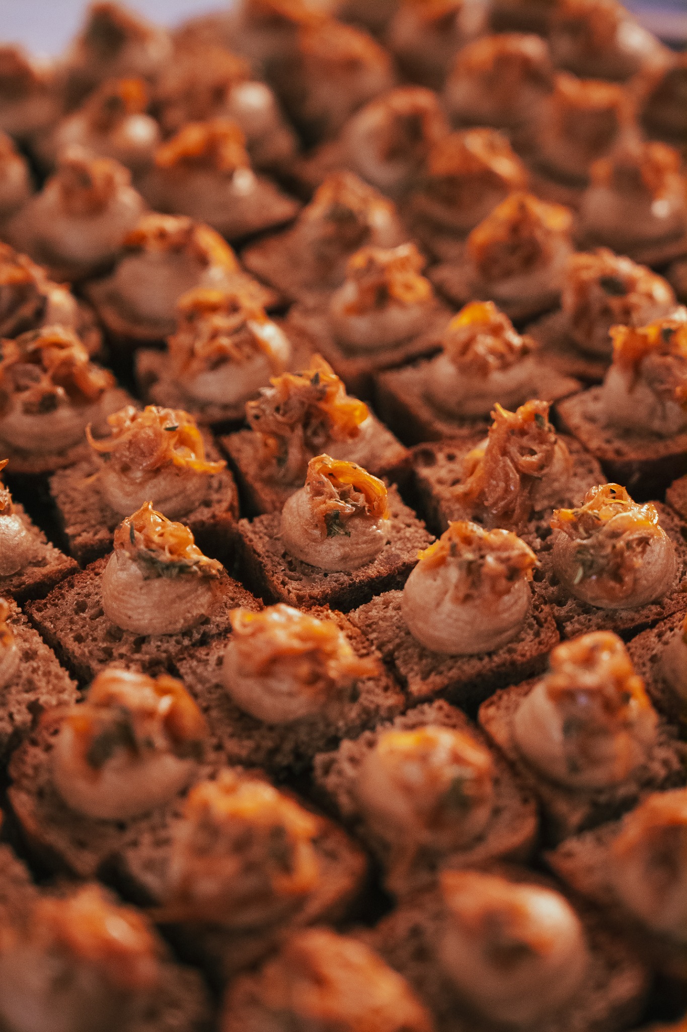 Apetyczne, równo ułożone kanapeczki na ciemnym pieczywie, udekorowane kremowym farszem i karmelizowaną cebulką, tworzące elegancką kompozycję cateringową.