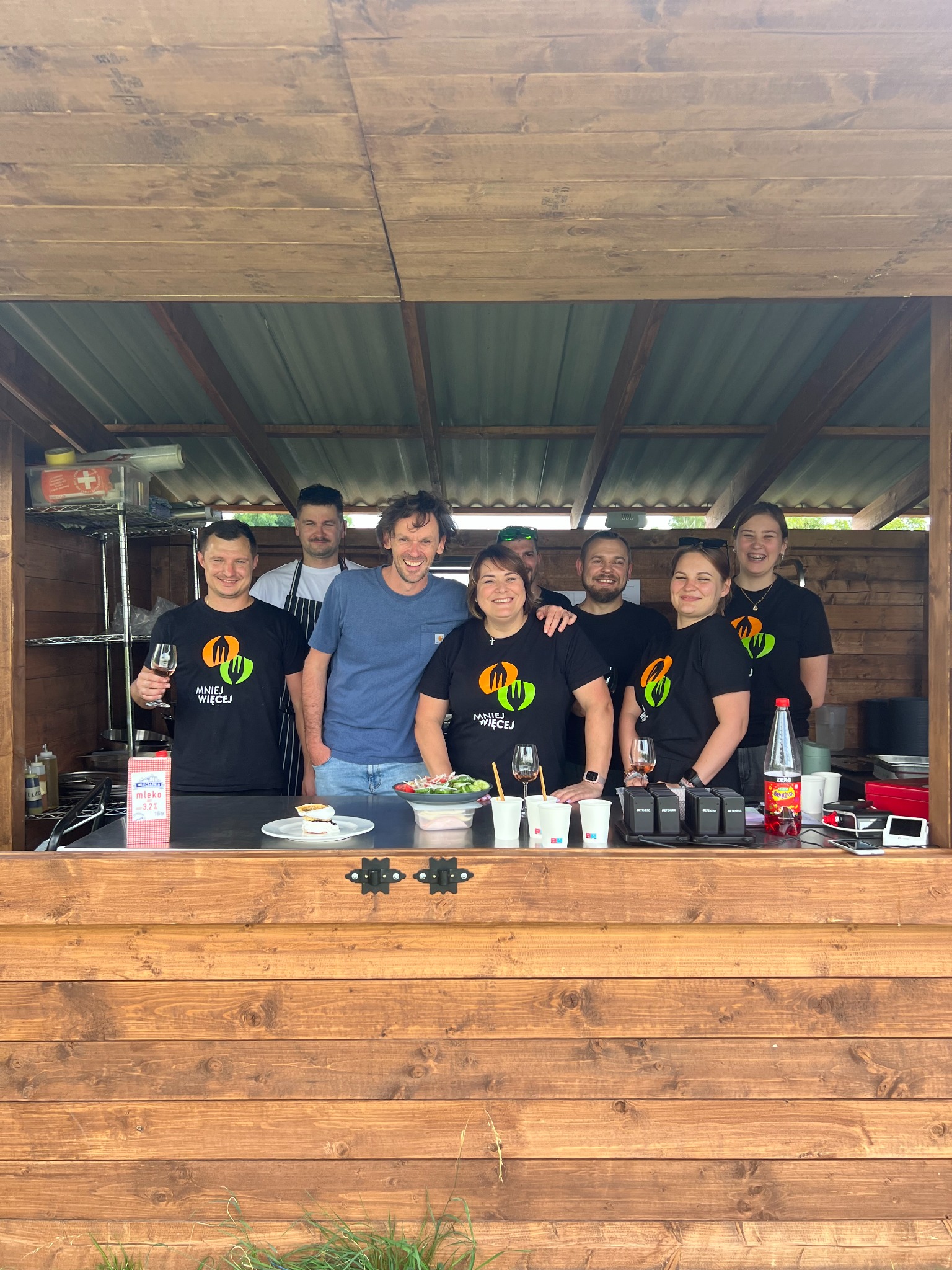 Zespół cateringowy w drewnianym barze plenerowym, prezentujący dania i napoje. Część osób w koszulkach z logo firmy, uśmiechnięci, w tle widoczny sprzęt gastronomiczny.