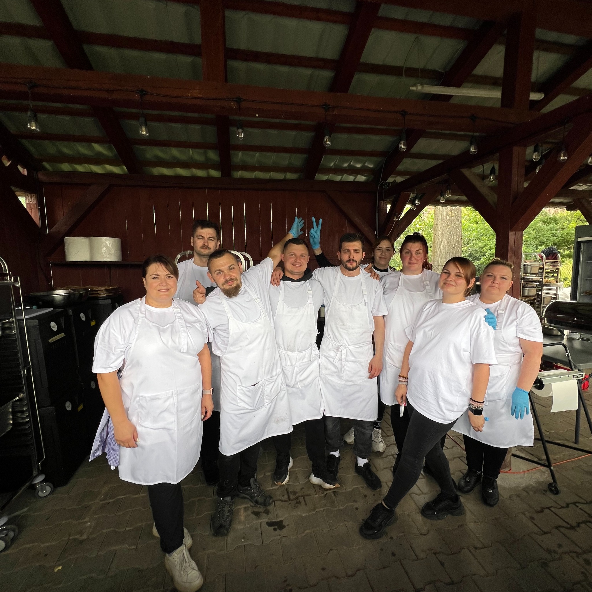 Zespół kucharzy w białych fartuchach i koszulkach pozuje na tle drewnianej konstrukcji z oświetleniem. Widoczny sprzęt gastronomiczny i kostka brukowa.
