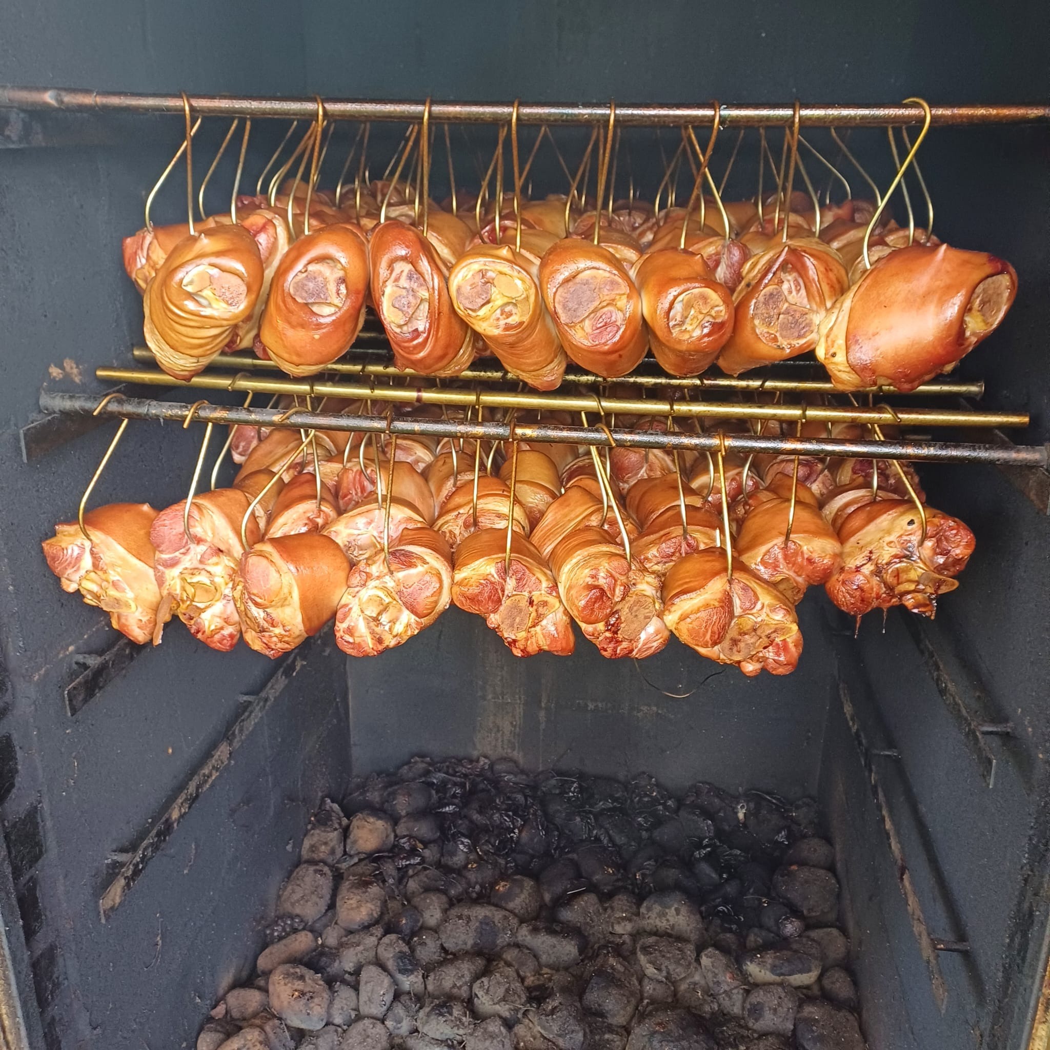 Wędzone golonki wieprzowe zawieszone na metalowych hakach wewnątrz wędzarni. Widoczne palenisko z węglem drzewnym. Obróbka termiczna mięs w gastronomii.