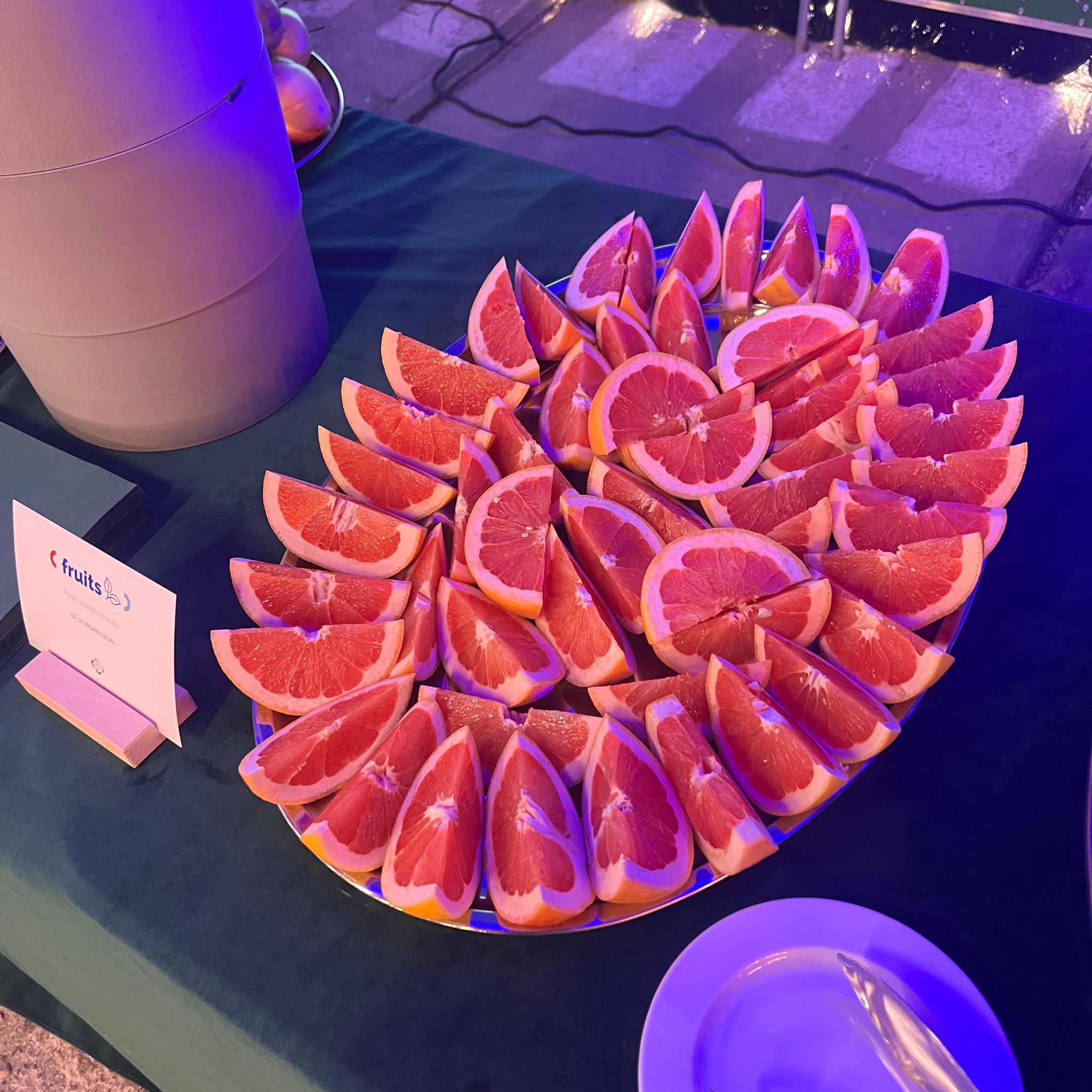 Talerz z pokrojonymi grejpfrutami na ciemnozielonym obrusie, obok dyspenser na napoje i tabliczka z napisem 'fruits'. Catering dietetyczny.