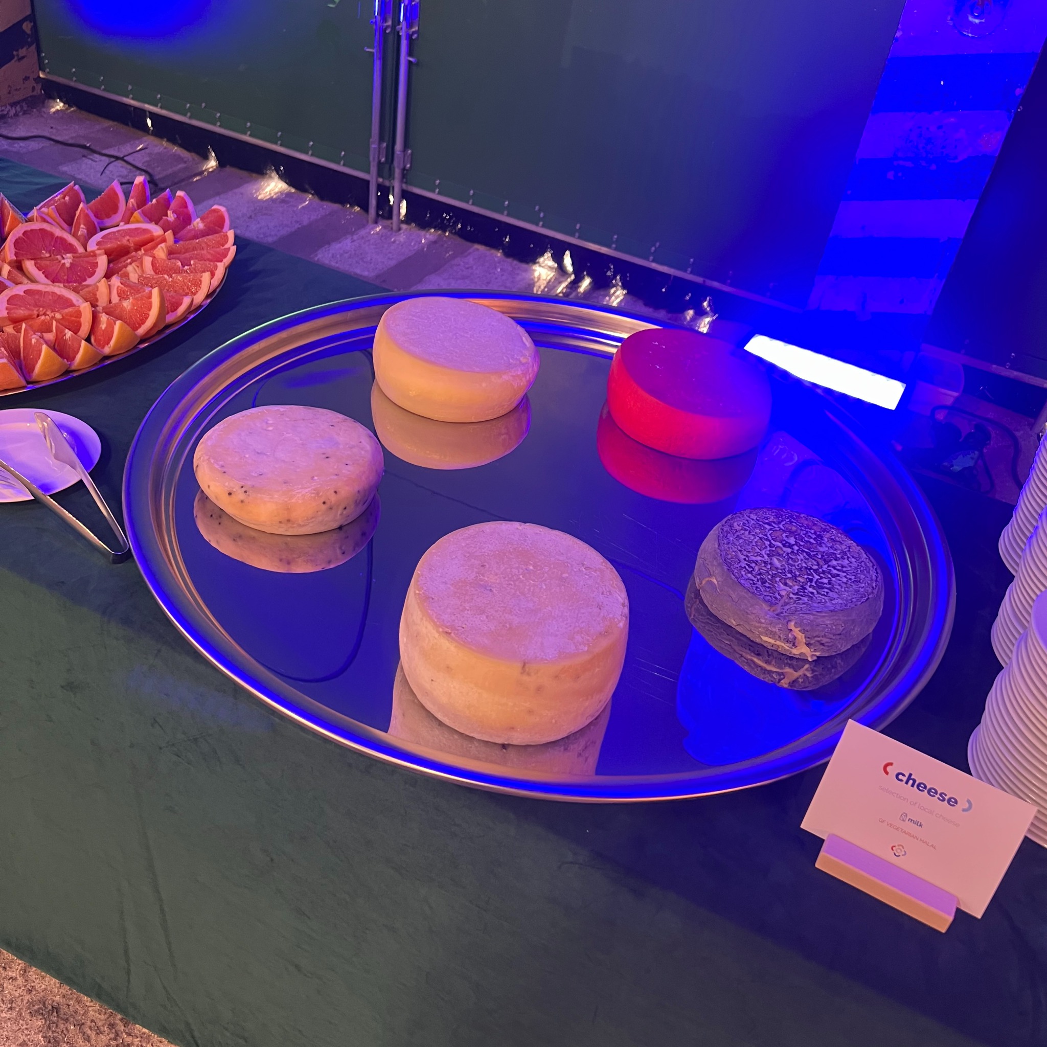 Srebrna taca z selekcją serów (różne rodzaje i kolory) oraz grejpfrutów na zielonym obrusie, oświetlona niebieskim światłem, z widoczną etykietą 'cheese'.