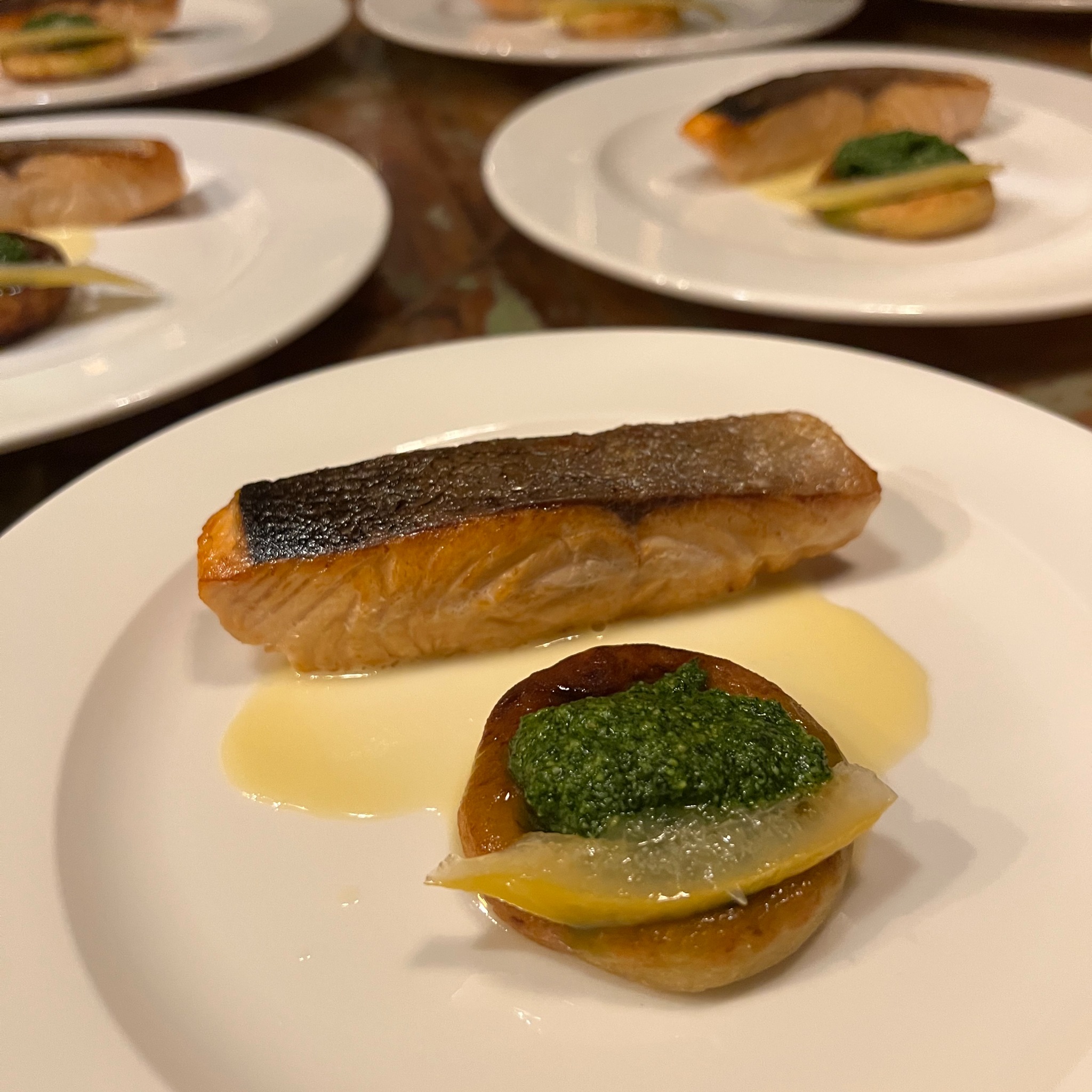 Elegancko podany łosoś z zielonym pesto na talerzu. Widoczne inne porcje, co sugeruje catering lub przyjęcie.