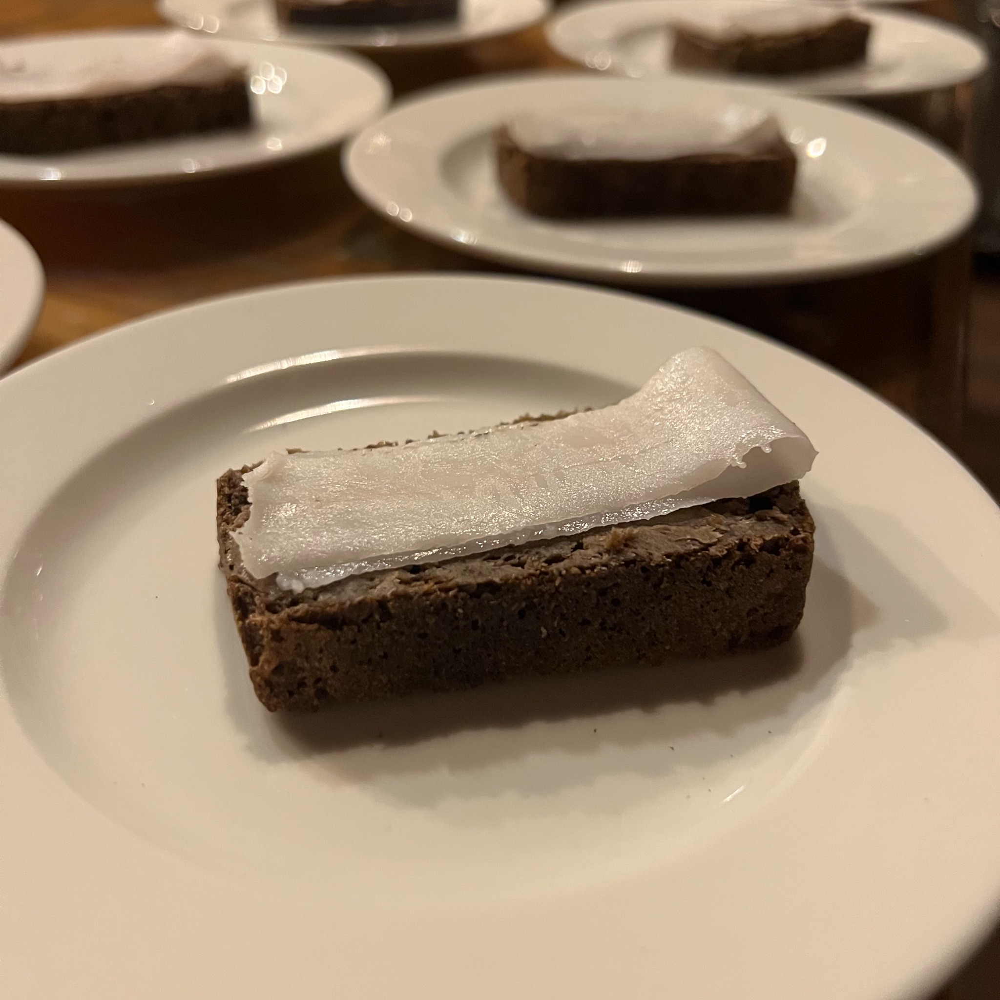 Przekąska: kromka ciemnego chleba z pastą i plasterkiem słoniny na białym talerzu. W tle kilka innych talerzy z podobnymi przekąskami.