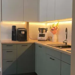 Nowoczesna kuchnia z oliwkowymi szafkami i drewnianym blatem. Oświetlenie LED podkreśla marmurowy backsplash i dodaje ciepła wnętrzu. Minimalistyczny design.