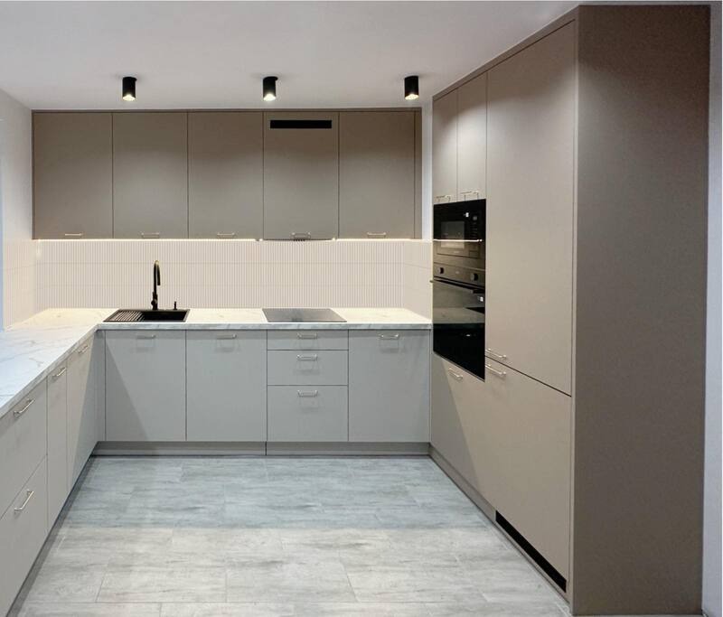 Jasna kuchnia z marmurowym blatem i czarną armaturą. Górne szafki w kolorze taupe, dolne szaro-beżowe. Nad blatem białe płytki z subtelnym wzorem.