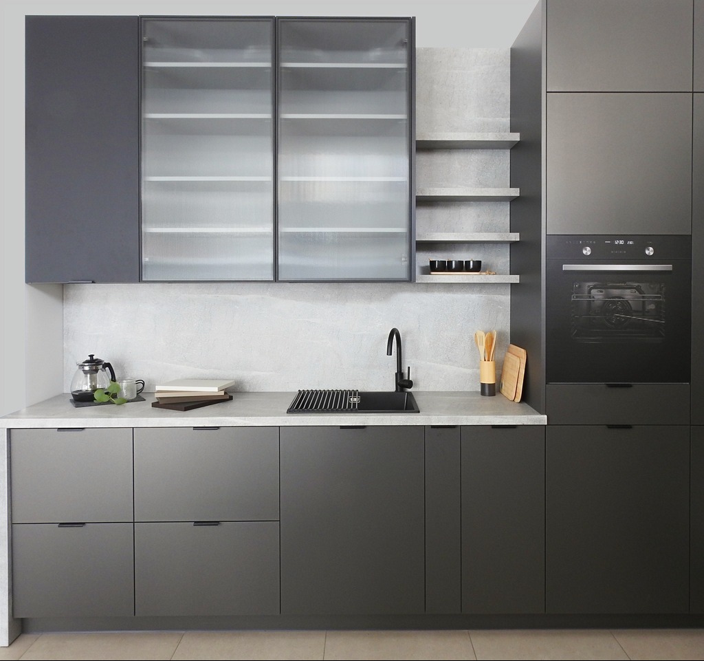 Nowoczesna kuchnia w odcieniach szarości z matowymi frontami, przeszklonymi szafkami górnymi i minimalistycznym oświetleniem. Uwagę zwraca czarny zlew i bateria.