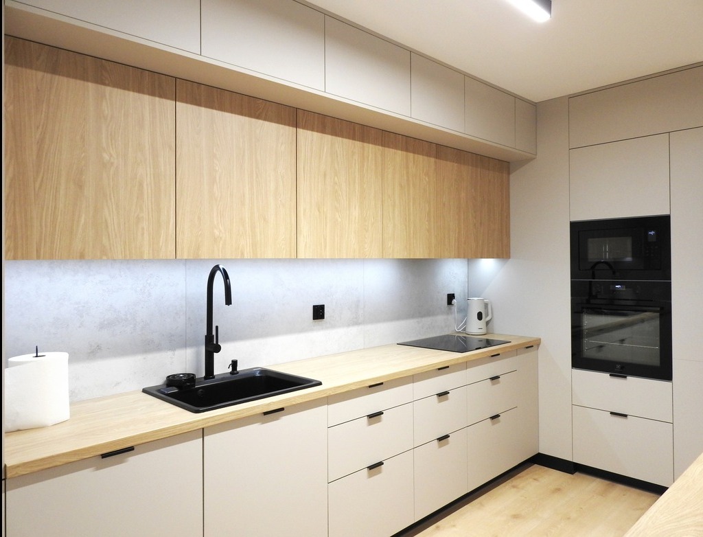 Nowoczesna kuchnia z drewnianymi górnymi szafkami i czarną armaturą. Jasne, minimalistyczne fronty dolnych szafek z dyskretnymi uchwytami.