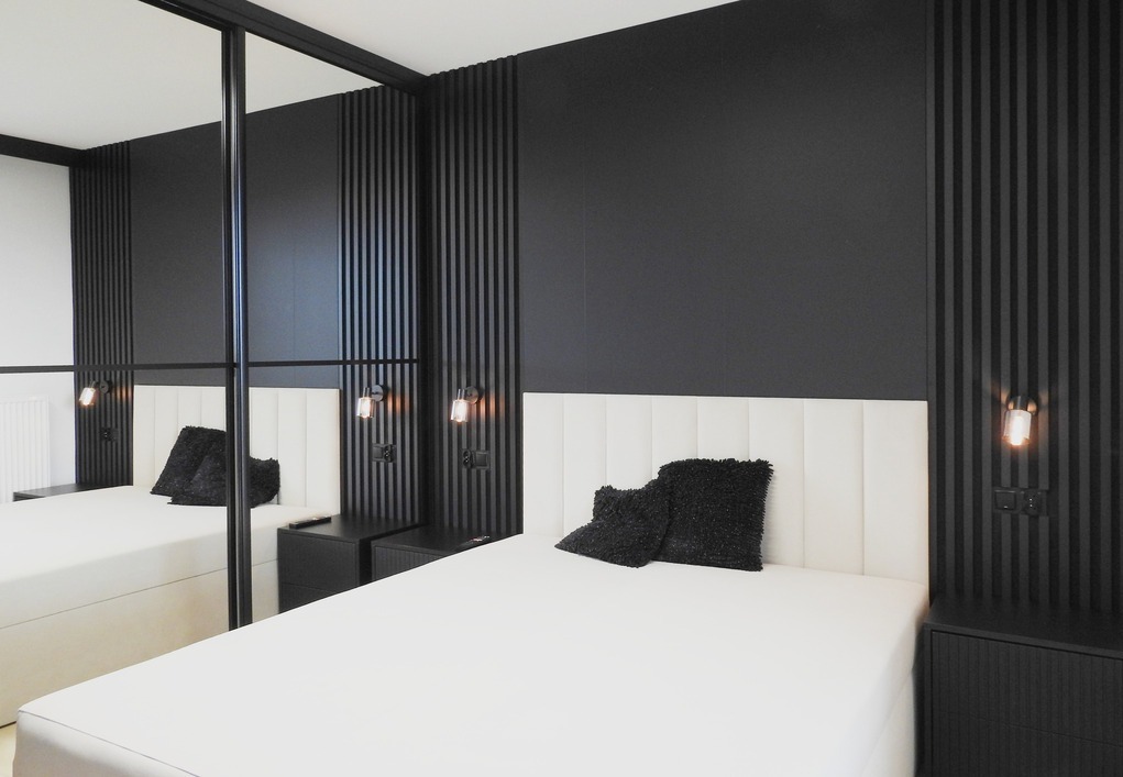 Nowoczesna sypialnia z łóżkiem, szafkami nocnymi i ścianą z lameli w kolorze czarnym. Lustrzane drzwi szafy odbijają wnętrze. Oświetlenie punktowe.