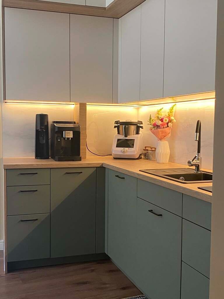 Nowoczesna kuchnia z oliwkowymi szafkami i drewnianym blatem. Oświetlenie LED podkreśla marmurowy backsplash i dodaje ciepła wnętrzu. Minimalistyczny design.