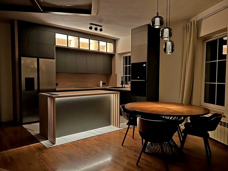 Nowoczesna kuchnia z wyspą i oświetleniem LED. Czarne szafki, drewniany blat i stół z krzesłami tworzą spójną całość. Ciepłe światło lamp wiszących.