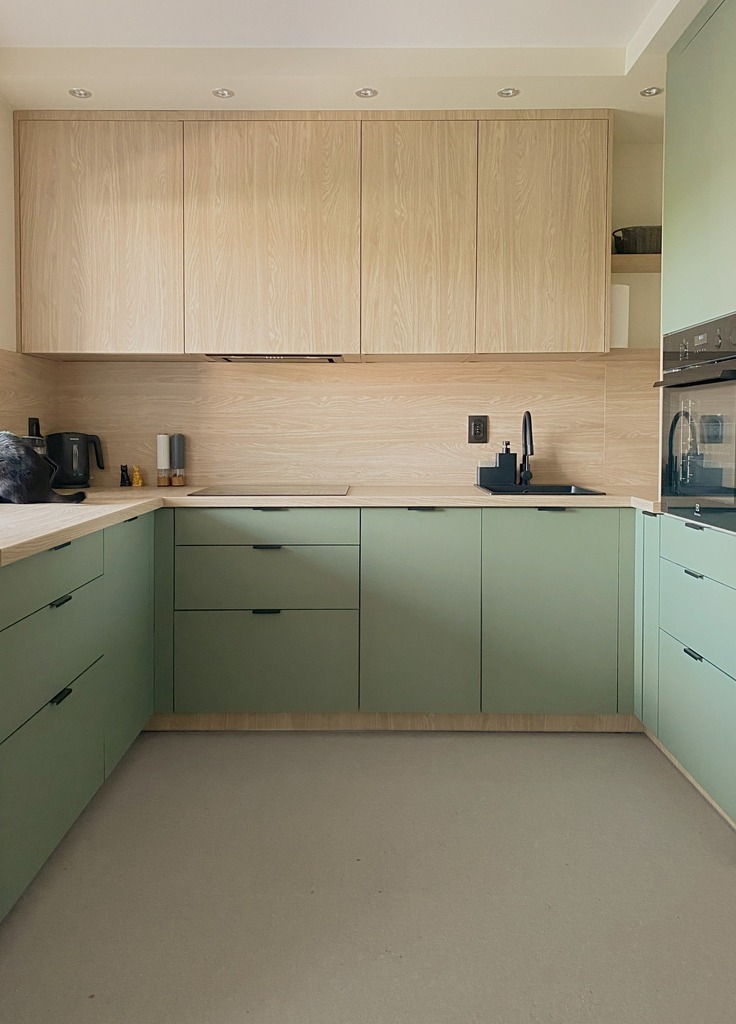 Jasna kuchnia w kształcie litery U z zielonymi szafkami i drewnianymi blatami. Górne szafki w kolorze drewna, czarne uchwyty i armatura. Minimalistyczny design.