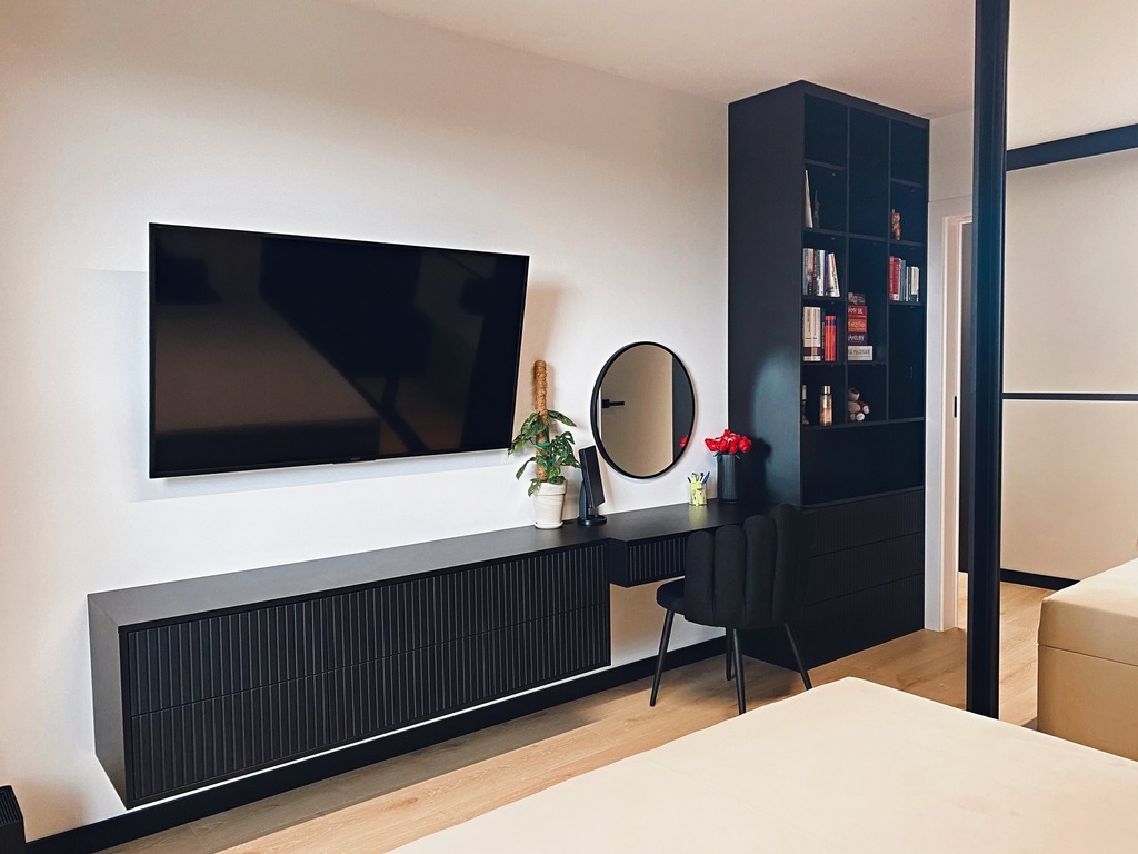 Czarna zabudowa meblowa w sypialni z biurkiem, szafką RTV i regałem. Minimalistyczny design, oświetlenie LED pod szafką, okrągłe lustro nad blatem. Nowoczesny styl.
