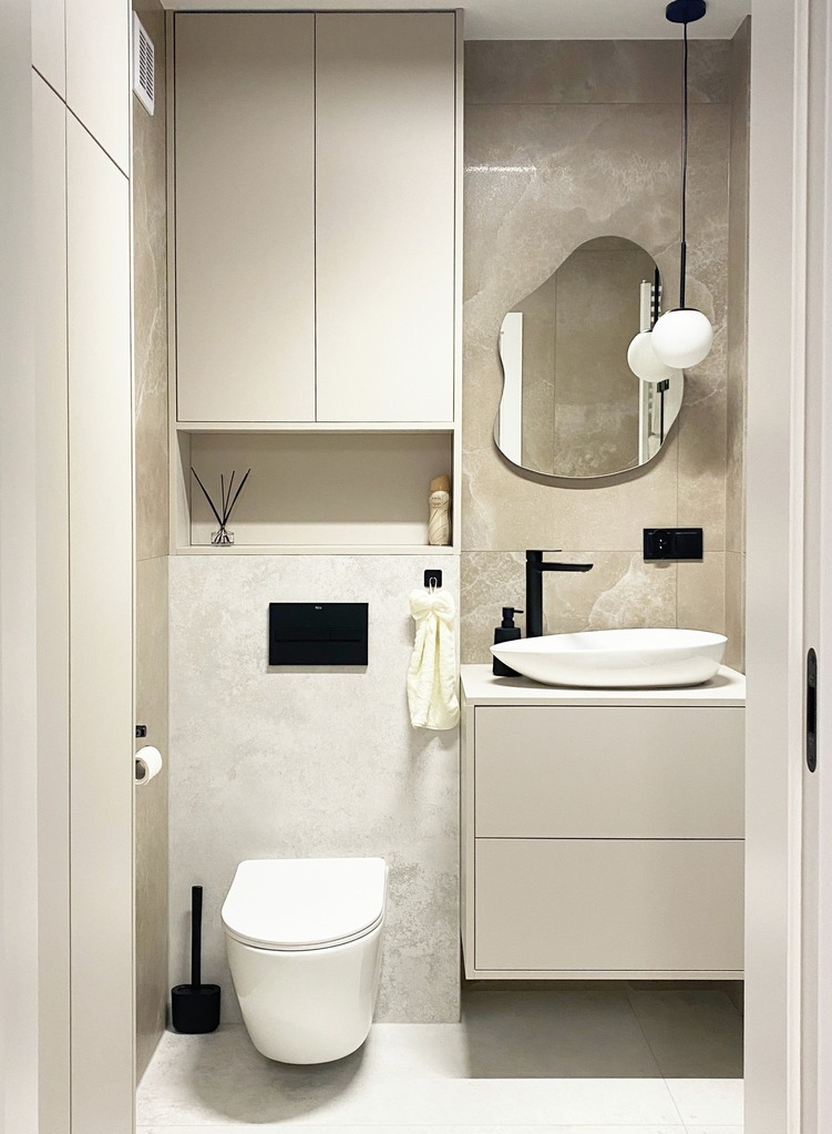 Nowoczesna łazienka w beżach: podwieszana toaleta, umywalka nablatowa z szafką, lustro o nieregularnym kształcie i szafka wisząca. Minimalistyczny design.