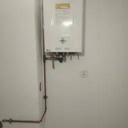 Biały piec gazowy dwufunkcyjny, zamontowany na ścianie z widocznymi miedzianymi rurami i zaworami, podłączony do instalacji. Widoczny gniazdko elektryczne.