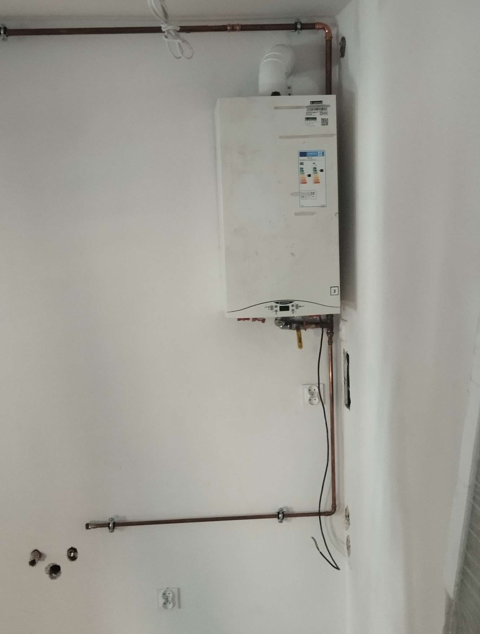 Biały piec gazowy Ariston zamontowany na ścianie z widocznymi miedzianymi rurami instalacji grzewczej i otworami w ścianie po starych instalacjach.