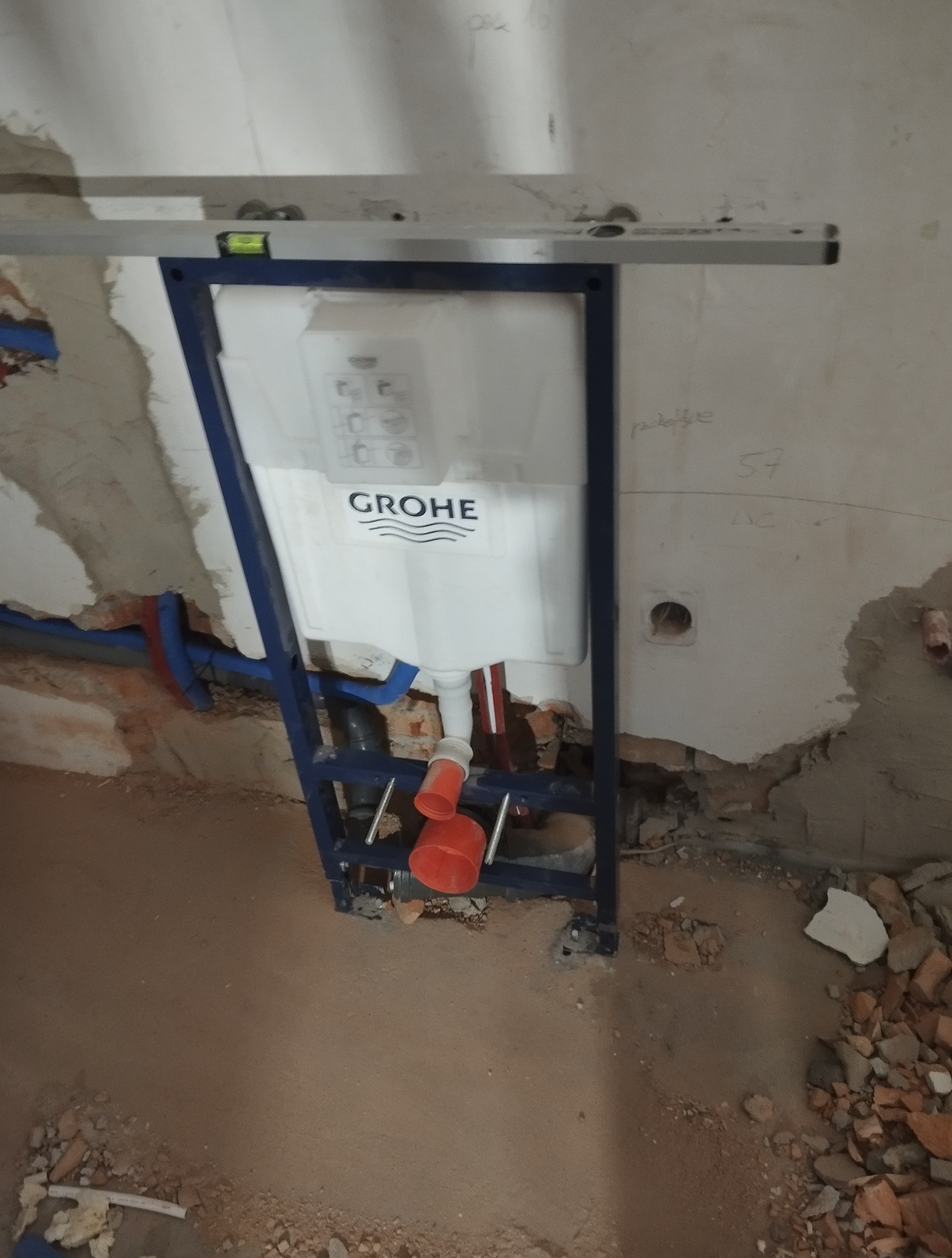 Stelaż podtynkowy Grohe z poziomnicą, widoczne przyłącza wodne i kanalizacyjne. Etap montażu w surowym stanie. Fragment ściany z instalacją i gruz na podłodze.