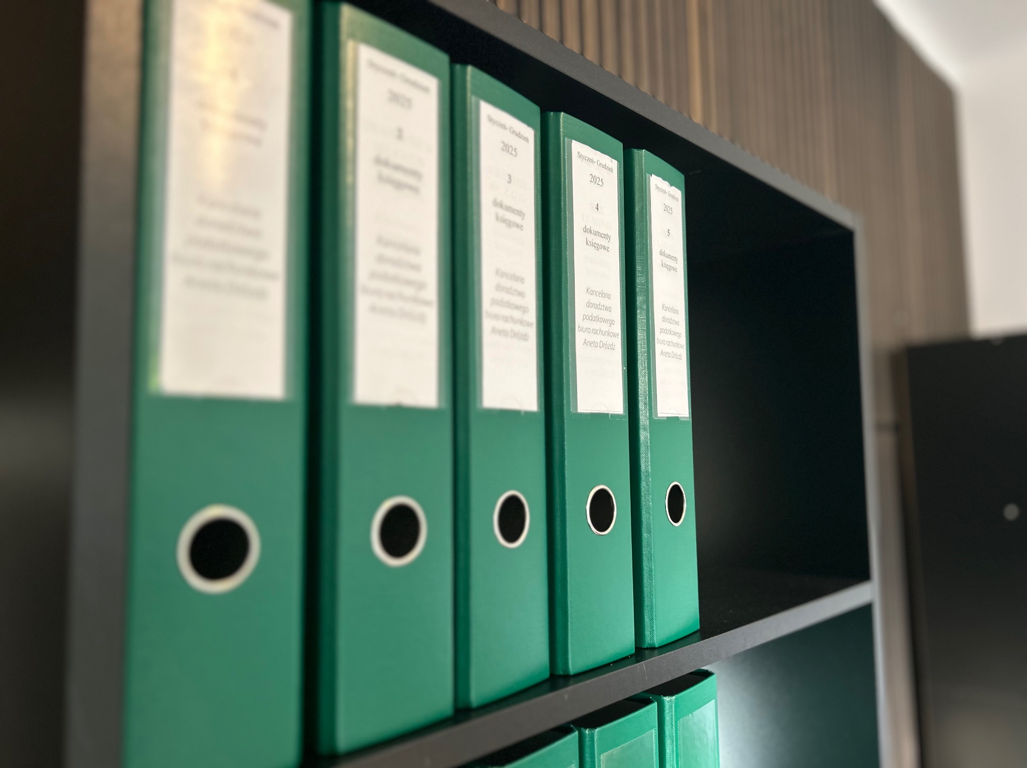 Rząd zielonych segregatorów z dokumentami księgowymi na półce, oznaczonych etykietami z nazwą kancelarii i rokiem 2025. Ujęcie z bliska, oświetlenie biurowe.