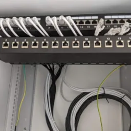 Panel krosowniczy z podłączonymi kablami sieciowymi w szafie rack, widoczne porty RJ45 i kable Ethernet w kolorach białym, szarym i czarnym.